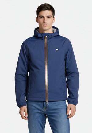 Man die een blauwe capuchonjas draagt met een witte en oranje gestreepte ritssluiting, gecombineerd met een blauwe spijkerbroek, staand tegen een witte achtergrond.