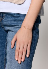 Złota bransoletka łańcuszkowa z małym zapięciem i ozdobnym charms'em, umieszczona na tle dżinsu i jasnej bluzki.