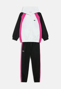 Trening alb-negru cu panouri laterale roz. Are o jachetă cu fermoar și pantaloni cu talie elastică. Include un logo mic verde pe jachetă.