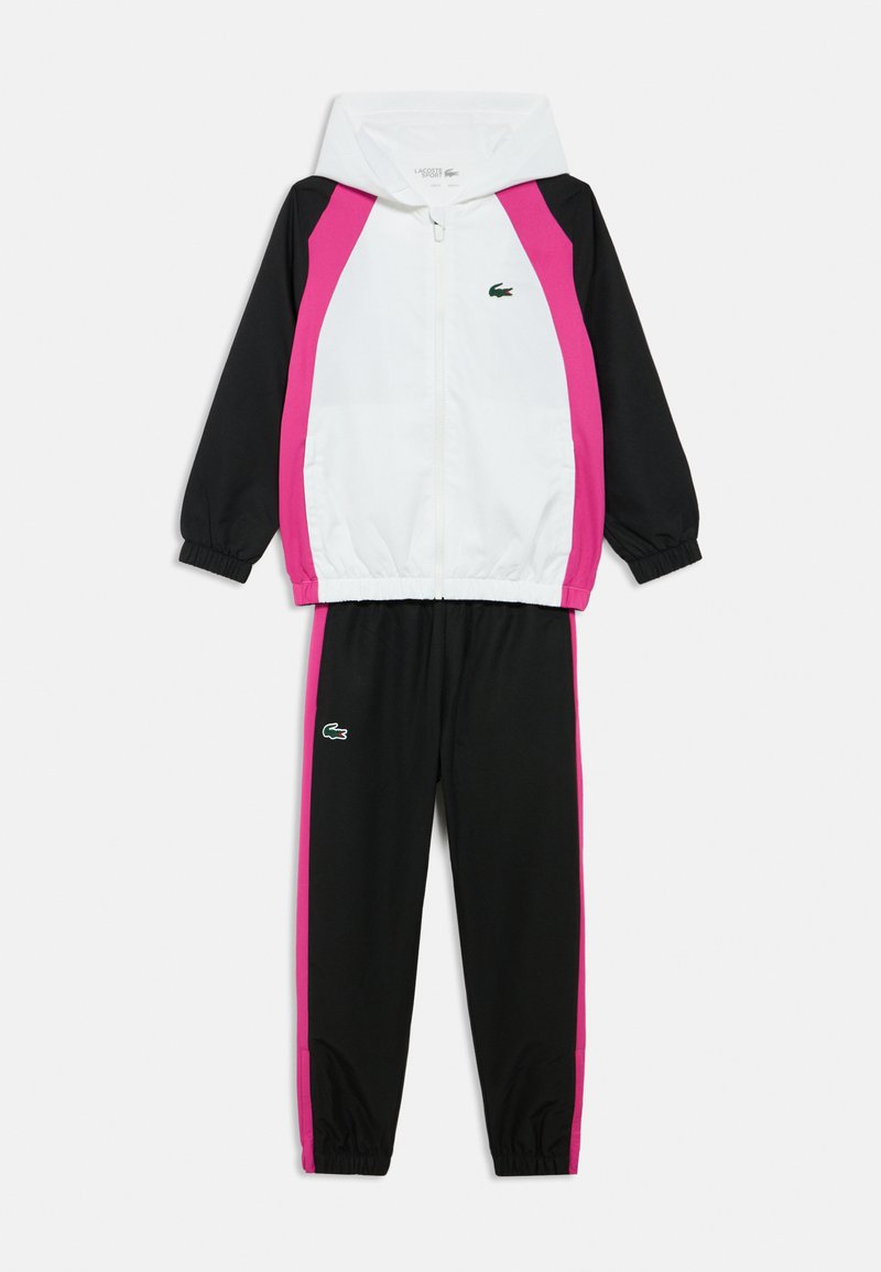 Zwart-wit trainingspak met roze zijpanelen. Bestaat uit een jas met ritssluiting en een broek met elastische tailleband. Bevat een klein groen logo op het jack.