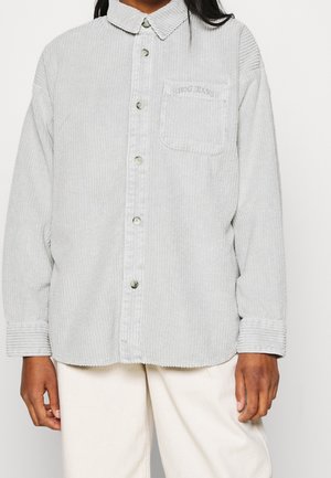 Personne portant une chemise en velours côtelé gris clair à boutons avec une poche poitrine étiquetée « BDG JEANS » et un pantalon écru.
