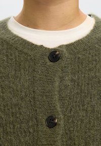 Zöld kötött kardigán bolyhos anyagból, V-nyakkivágással, két fekete gombbal és bordázott textúrával. Alatta fehér ing van.