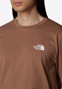 Bruine katoenen t-shirt met een ronde halslijn en korte mouwen. Op de linkerborst staat een wit logo van The North Face in een gebogen ontwerp.