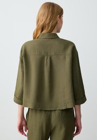 Oliwkowa, lekka lniana kurtka o nieco luźnym kroju, z złożonymi mankietami i szczegółem w postaci plisek z tyłu. Minimalistyczny design, bez widocznych zapięć.