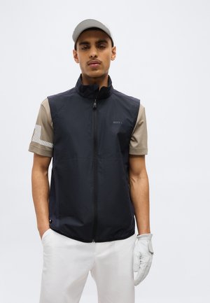 Noor mees kannab musta tõmbekinnitusega vesti beeži särgi üle, paremal käel valget golfikinda, valgeid pükse ja helehalli nokamütsi, seisab käed taskutes.