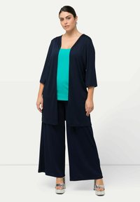 Veste kimono navy ouverte avec des manches 3/4, associée à un top en teinte teal et un pantalon large navy, complété par des chaussures à plateforme argentées.