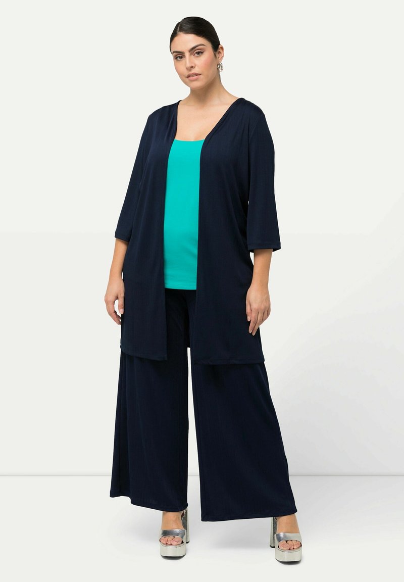 Veste kimono navy ouverte avec des manches 3/4, associée à un top en teinte teal et un pantalon large navy, complété par des chaussures à plateforme argentées.