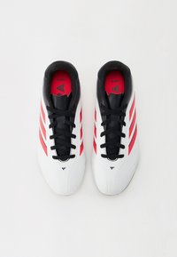 Zapatos de fútbol en blanco y negro, con tres franjas rojas, diseño con cordones y una textura suave. Punta redondeada y silueta baja.