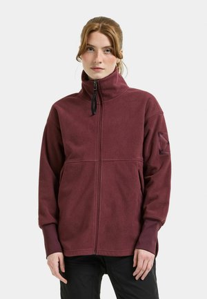 Frau trägt eine weinrote Fleecejacke mit hohem Kragen und Daumenlöchern an den Ärmelbündchen sowie schwarze Hose, steht vor einem einfarbigen Hintergrund.