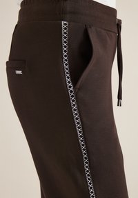 Pantalon marron foncé avec poches latérales, bande décorative blanche à motifs le long du bord de la poche, et taille ajustable à cordon avec embouts métalliques.