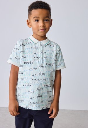 Băiat tânăr cu păr scurt și creț, purtând un tricou polo alb cu fermoar, modelat cu textul „Ted Baker” și pantaloni închiși la culoare, stând în picioare pe un fundal simplu.