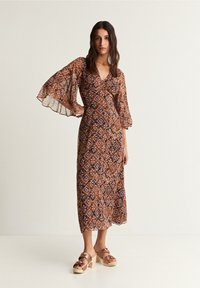 Maxi jurk met bell mouwen in bruin, met een geometrisch patroon in tinten van crème en paars. Gepaarde met lichtbruine platform sandalen.