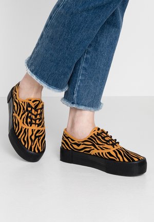 Pieds portant des baskets compensées rayées tigre orange et noir associées à un jean en denim bleu à ourlet effiloché sur un sol blanc.