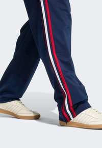 Námornícke tepláky s bielymi a červenými pruhmi po boku, so zipsom na členku, v kombinácii s bielymi športovými topánkami a textúrovanou podrážkou.