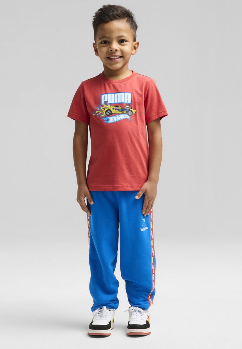 Camiseta de algodón roja con un gráfico azul de "PUMA" y un coche, combinada con pantalones de chándal azules con franjas laterales amarillas y rojas. Zapatillas en blanco y negro.