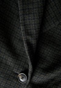 Tessuto di blazer grigio scuro con una sottile texture, caratterizzato da un bottone metallico con quattro fori e testo inciso. Il tessuto appare morbido e leggermente peloso.