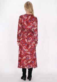 Langes rotes Kleid mit Paisley- und Blumenmuster, langen Ärmeln, taillierter Taille und fließendem Silhouette. Getragen mit schwarzen kniehohen Stiefeln.