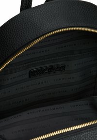 Mochila de cuero texturizado negro con cremallera dorada. El interior cuenta con un forro negro con la marca "TOMMY HILFIGER" repetida en un patrón de rayas.