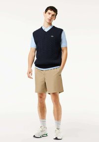 Marinblå kabelstickad väst över ljusblå polo, i kombination med tanfärgade shorts. Vita sneakers med gröna accenter och randiga strumpor.