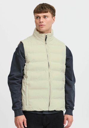 Hellbeige, wattierte Weste mit hohem Kragen, Reißverschluss und seitlichen Taschen. Über einem dunklen Langarmshirt getragen. Glatte Textur.