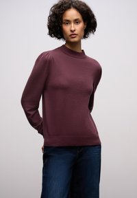 Bordeauxfarbener Pullover mit hohem Kragen, langen Ärmeln und geripptem Saum. Weiche Textur mit dezentem Glanz. Kombiniert mit dunkelblauen Jeans.