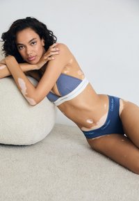 Bralet din material côtelat Calvin Klein, de culoare albastră, cu bandă albă, asortat cu chiloți albaștri cu logo. Modelul pozează pe o suprafață moale bej.