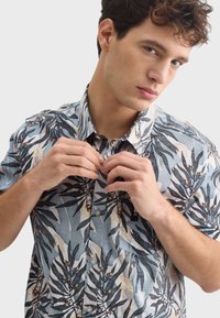 Košeľa na gombíky s krátkym rukávom z svetlomodrej látky, ktorá obsahuje čierny a zelený vzor listov, s jemnými doplnkami oranžových kvetov a štítkom na boku.