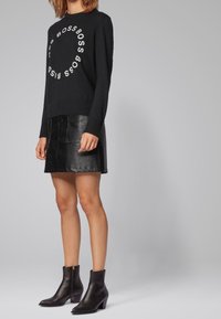 Schwarzer Strickpullover mit rundem weißen Logo, kombiniert mit einem schwarzen Lederrock und Stiefeletten, die ein schlankes Design präsentieren.