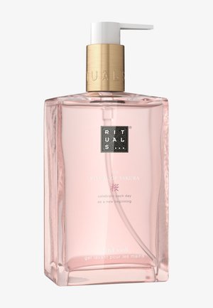 Botella rectangular transparente con un líquido rosa. Cuenta con una tapa de color dorado y un dispensador de bomba. El texto incluye "The Ritual of Sakura" y "lavado de manos".