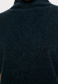 Gros plan d'un pull à col roulé tricoté texturé vert foncé porté par une personne, montrant le détail du tissu et le col montant.