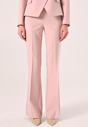 Kobieta ubrana w jasnoróżowe, dopasowane spodnie i pasującą marynarkę, beżowe buty w szpic, stojąca na tle jednolitego tła.