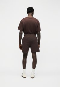 Άνδρας που στέκεται γυρισμένος προς τα πίσω, φοράει σκούρο καφέ μπλουζάκι και ταιριαστά σορτς, λευκές κάλτσες και μπεζ αθλητικά παπούτσια σε ένα απλό λευκό φόντο.