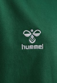 Grön bomullstyg med vitt broderat logotyp med stiliserad bi-design och texten "hummel" nedanför. Slät textur, närbild.