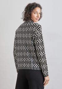 Femme aux cheveux courts et bouclés portant un pull à motifs géométriques noirs et blancs, regardant par-dessus son épaule sur un fond uni.