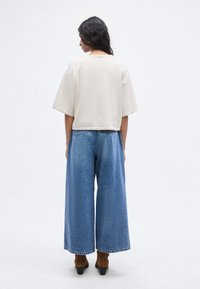 Μπεζ cropped μπλουζάκι με κοντά μανίκια, συνδυασμένο με μπλε τζιν παντελόνι φαρδιάς γραμμής. Διαθέτει ακατέργαστο στρίφωμα και άνετη εφαρμογή. Καφέ μποτάκια μέχρι τον αστράγαλο.