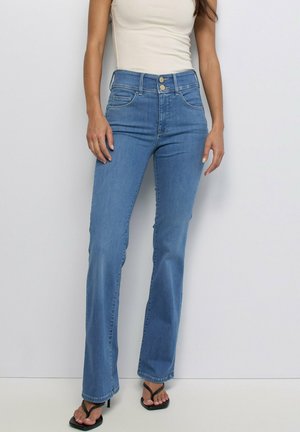 Femme portant un jean bleu taille haute avec double boutons, un haut blanc sans manches et des sandales à talons noires, debout devant un fond uni.