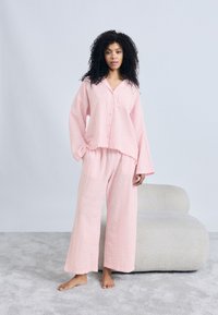 Σετ πιτζάμες με ροζ καρό σχέδιο που περιλαμβάνει πουκάμισο με κουμπιά και μακριά μανίκια, καθώς και φαρδιά παντελόνια, κατασκευασμένα από μαλακό υφασμάτινο υλικό.