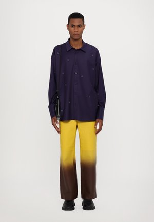 Paarse overhem shirt met ronde borduursels, gecombineerd met gele broek met een bruine gradient. Zwarte schoenen en een kleine zwarte schoudertas.