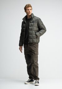 Gepolsterte olivgrüne Jacke mit hohem Kragen und zwei Seitentaschen, kombiniert mit dunklen Cargo-Hosen und schwarzen Sneakers. Glatte Textur, gesteppter Design.