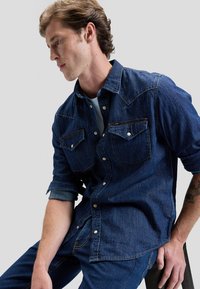 Vyras su susukti rankovėmis, apsirengęs tamsiai mėlyna džinsine palaidine ir džinsais, sėdintis ir viena ranka remiantis į šlaunį priešai paprastą foną.