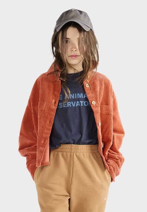 Orangene Cord-Hemd, marineblaues T-Shirt mit Text, beigen Hosen mit elastischem Bund und grauem Cap. Besitzt einen lässigen Schnitt und entspannten Stil.