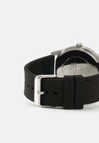 Bracelet de montre en silicone noir avec une boucle en métal argenté, présentant une texture lisse et un design perforé sur le côté interne.