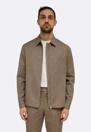 Uomo che indossa una giacca taupe con zip e tasca sul petto sopra una camicia bianca, abbinata a pantaloni taupe coordinati, in piedi davanti a uno sfondo semplice.