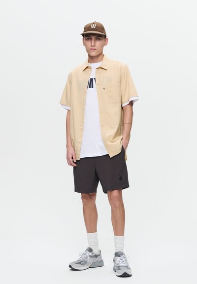 Chemise à manches courtes beige à boutons sur t-shirt blanc, shorts noirs, baskets grises, chaussettes blanches et casquette de baseball marron avec logo.