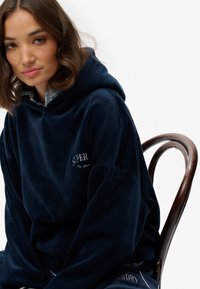 Sudadera de pana color azul marino con capucha grande, bolsillo frontal y logo de marca bordado. Modelo sentado en una silla de madera.