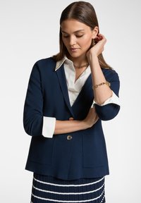 Blazer blu navy con una finitura testurizzata, bottoni dorati e tasche applicate, abbinato a una camicia bianca e a una gonna plissettata a righe.