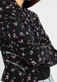 Blazer noir à motifs floraux avec de petites fleurs roses, blanches et bleues. Il présente des fermetures éclair argentées sur les poches latérales et un tissu texturé.