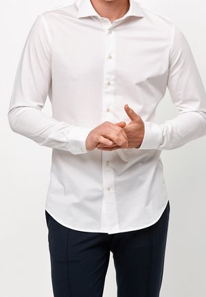 Homme ajustant les poignets d'une chemise blanche à manches longues boutonnée, portant un pantalon foncé, sur un fond clair uni.