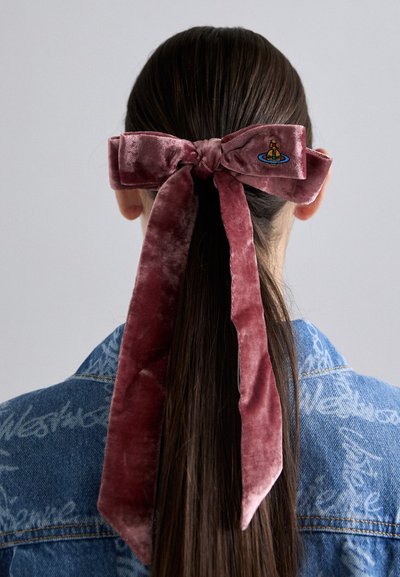 Sarkana rozā velūra matu lente ar lielu mezglu un garām astēm, ar nelielu izšuvumu vienā pusē, nēsāta kopā ar džinsu jaku.