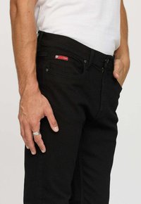 Homme portant un jean noir et une chemise blanche, avec une main reposant à ses côtés et l'autre dans sa poche, montrant une bague en argent.
